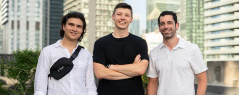 Three men (left man is wearing a white button-up shirt with a cross-bag, middle man is crossing his arms and wearing black shirt with grey pants, and the right man is wearing a white polo shirt) posing in front of skyscrapers