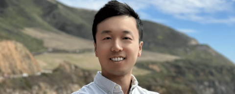 An Asian man in a white collar up shirt posing in front of mountains. in the right bottom corner you can see glimpses of a lake/sea