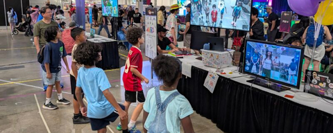 Group of children standing in a straight horizontal line and playing a soccer-like game. They are standing in front of a large tv screen and a laptop screen, which depict a VR game where the user is kicking a soccer ball 