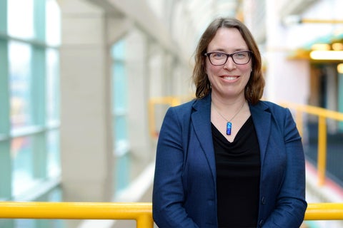 A brunette donning glasses, midnight-blue blazer, black shirt and a blue necklace posing in an open space. there are yellow bars in the background