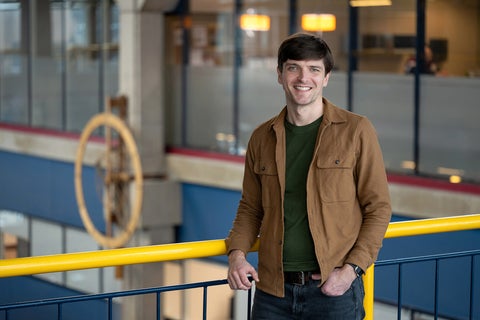 A person stands casually against a railing in an indoor space, wearing a brown jacket over a green shirt and dark jeans. A large wooden clock is visible in the background.