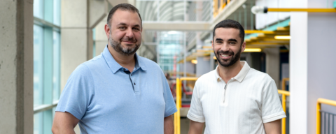 Two men (left person wearing a baby blue collaraed shirt with jeans and the right person wearing a white collared shirt with light grey pants) posing in front of a yellow rail