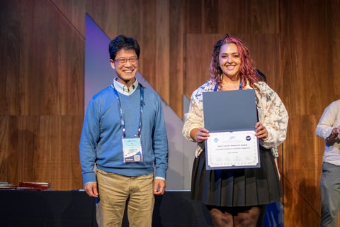 An Asian man in blue standing next to a Middle Eastern woman wearing a black dress and a white lacey jacket. She is holding her award
