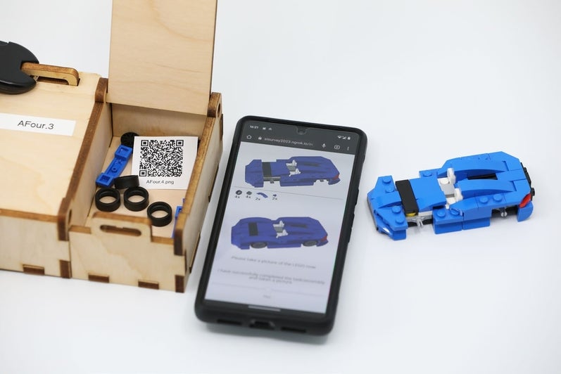 The fourth box is unlocked, showing a QR code and lego pieces. Next to the box are a phone showing online instructors and a built lego car