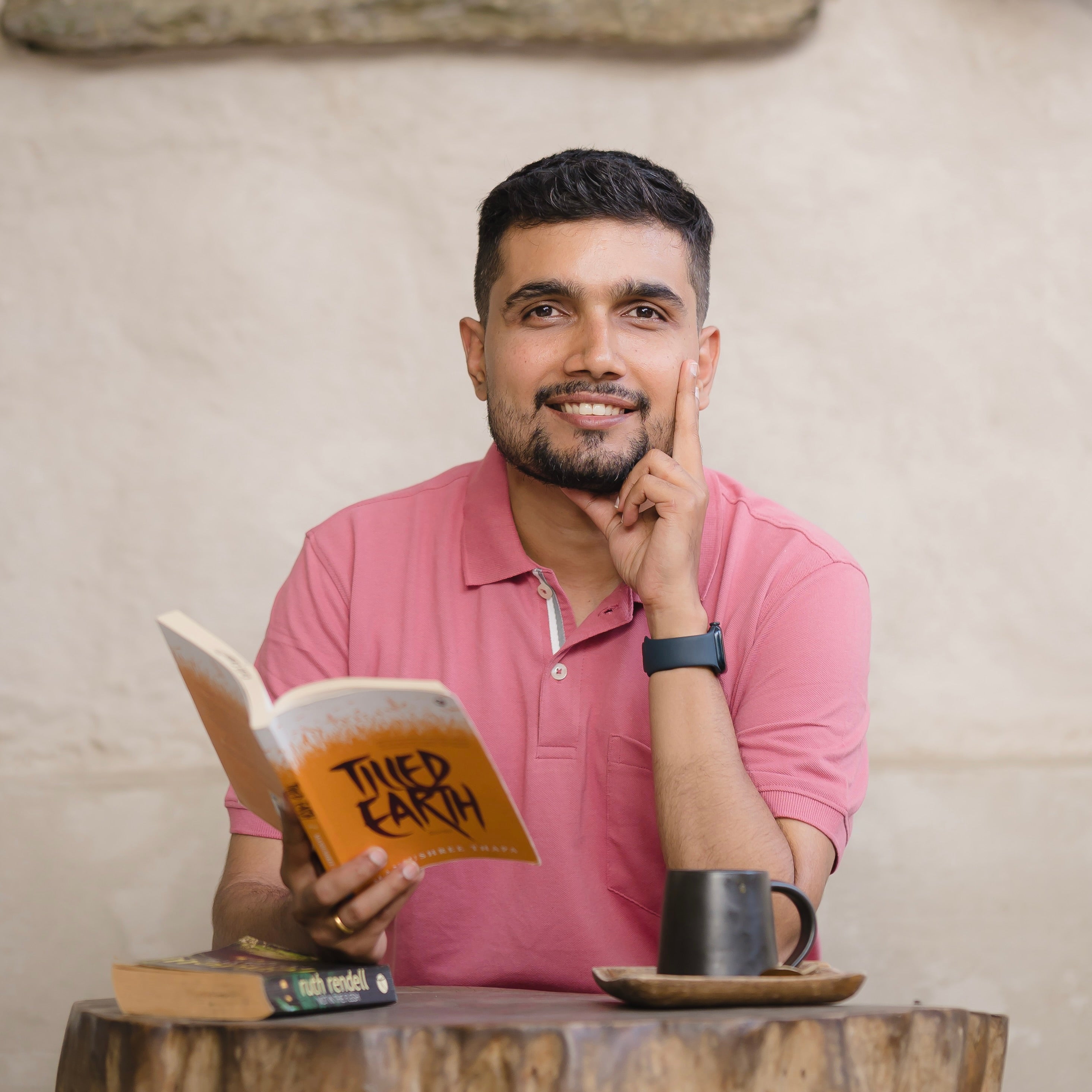 A South Asian male wearing a pink collared t-shirt. His right hand is holding his chin while his left hand is holding a orange-white book titled "Tilled Earth".