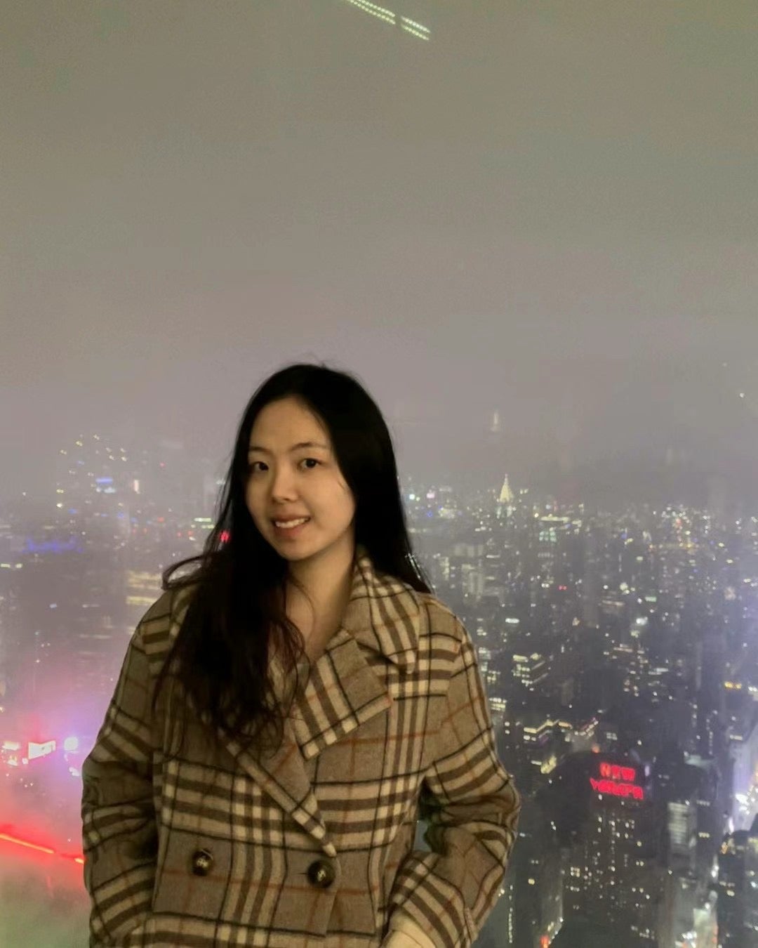 Photo of an Asian woman wearing a checked jacket standing aganist a large window. Behind her is a bunch of towers, taken at night