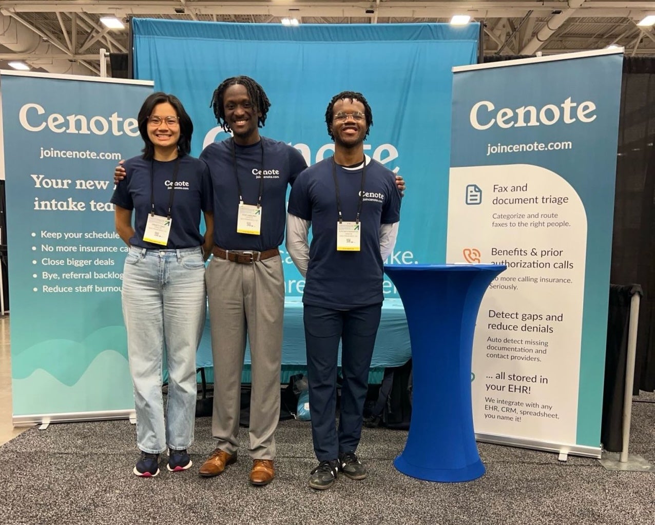 Three individuals stand in front of a Cenote booth with a blue backdrop and signage. They wear matching navy shirts and are positioned beside a blue table.