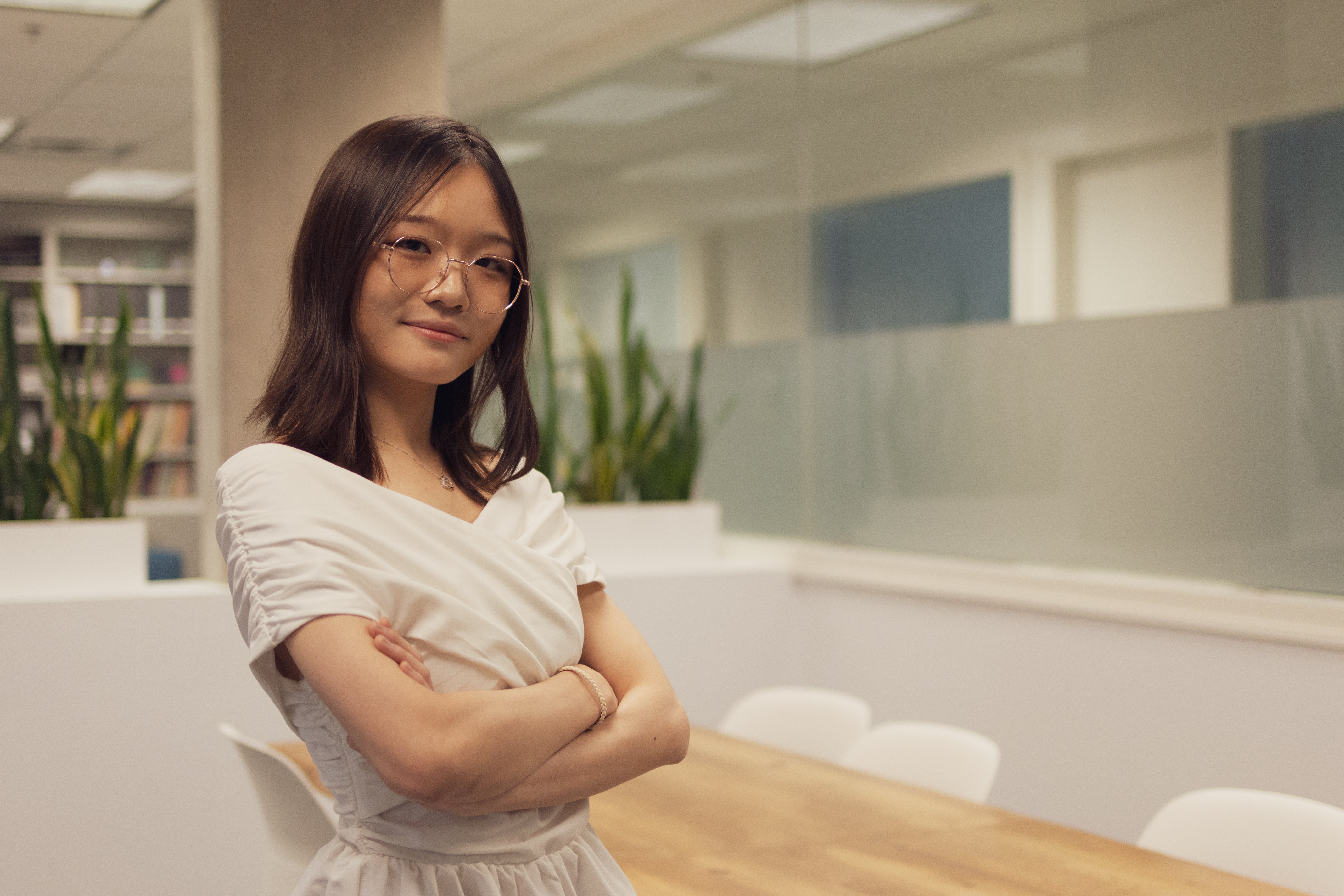 Photo of an Asian girl wearing glasses in a criss-cross off-the-shoulder top. She is crossing her arms and posing in a bright room. There's a desk behind her.