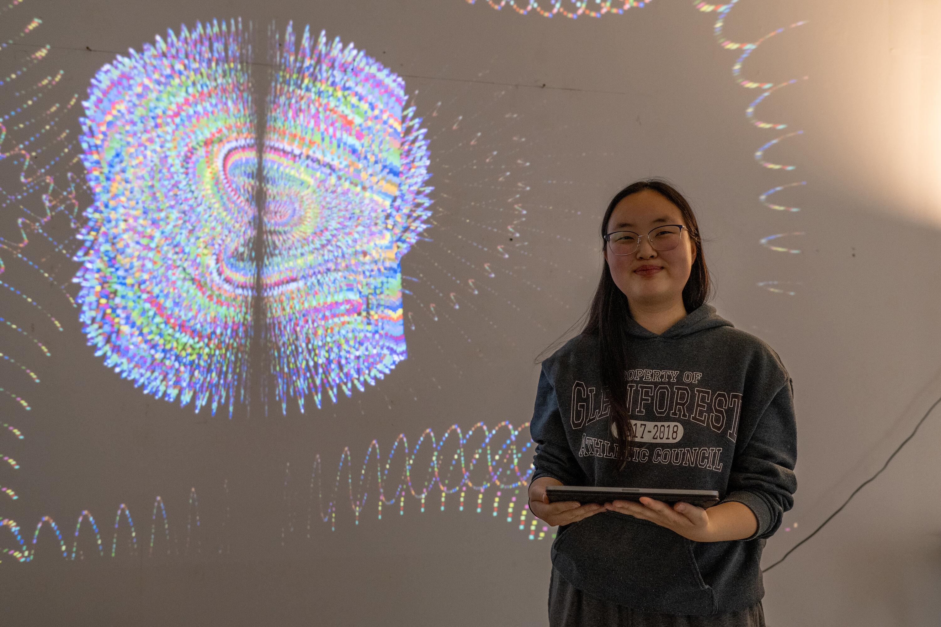 Student Hannah Choi posing in front of her installation Goodbye: where a blackscreen displays an interactive and colourful abstract artwork