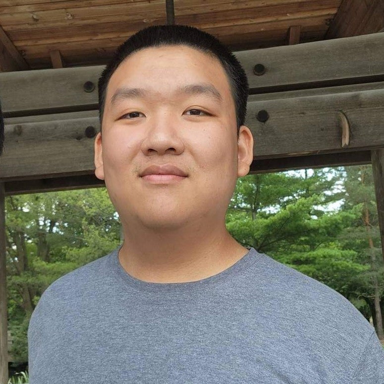 An Asian man wearing a blue-grey sweater posing in nature. He appears to be in a cabin