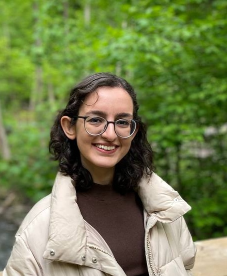 photo of an Iranian woman wearing glasses, a brown shirt and white puffer jacket. She is posing in a forest