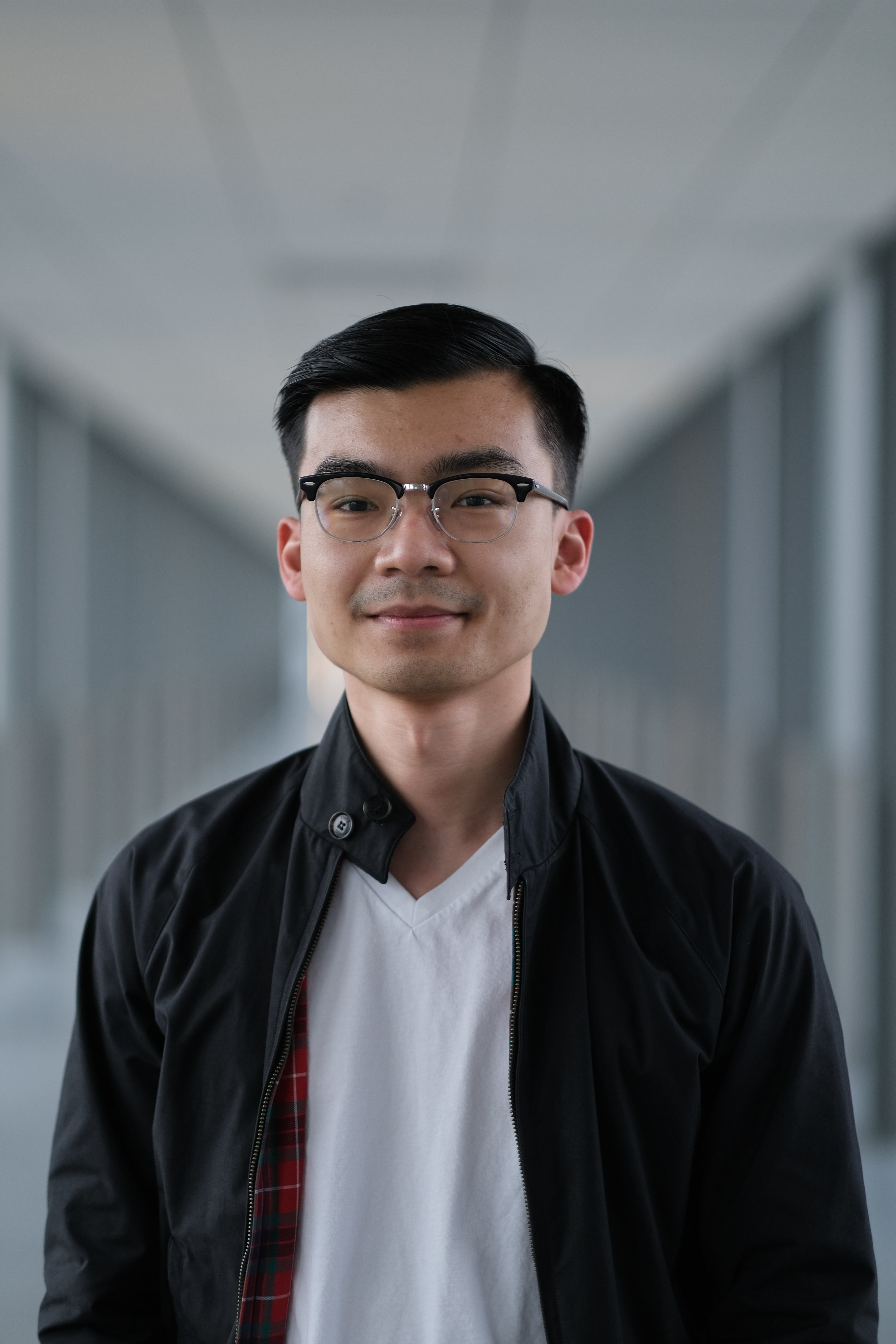 An Asian man wearing aviator-glasses, a black jacket and white shirt. He is posing inside of the E7 bridge hallway