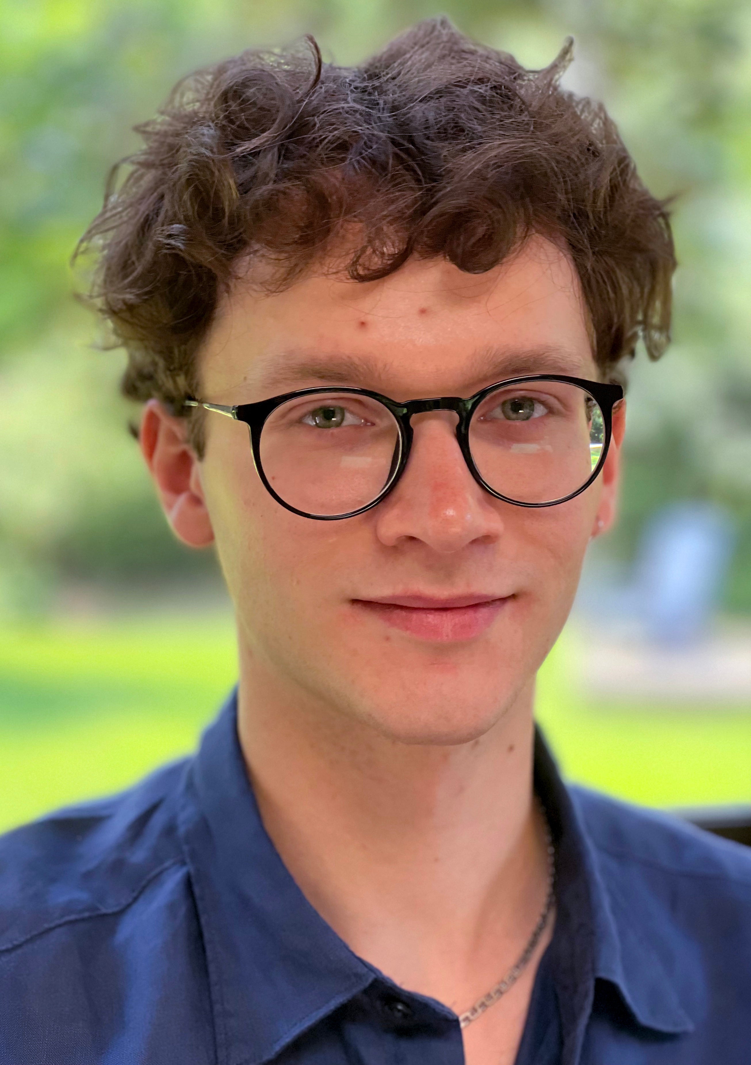 photo of a white man wearing black-rimmed glasses and a dark blue collar shirt. Photo taken outside (blurry)