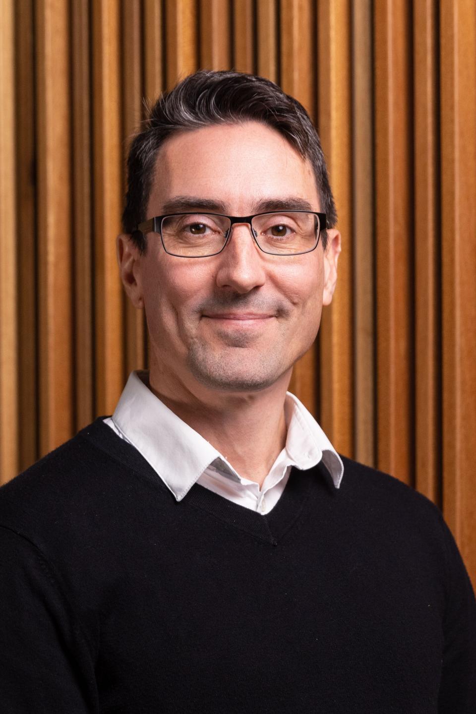 An european man wearing a black sweater on top of a white collared shirt posing aganist a wood-coloured accordion-looking wall. He is wearing rectangular glasses