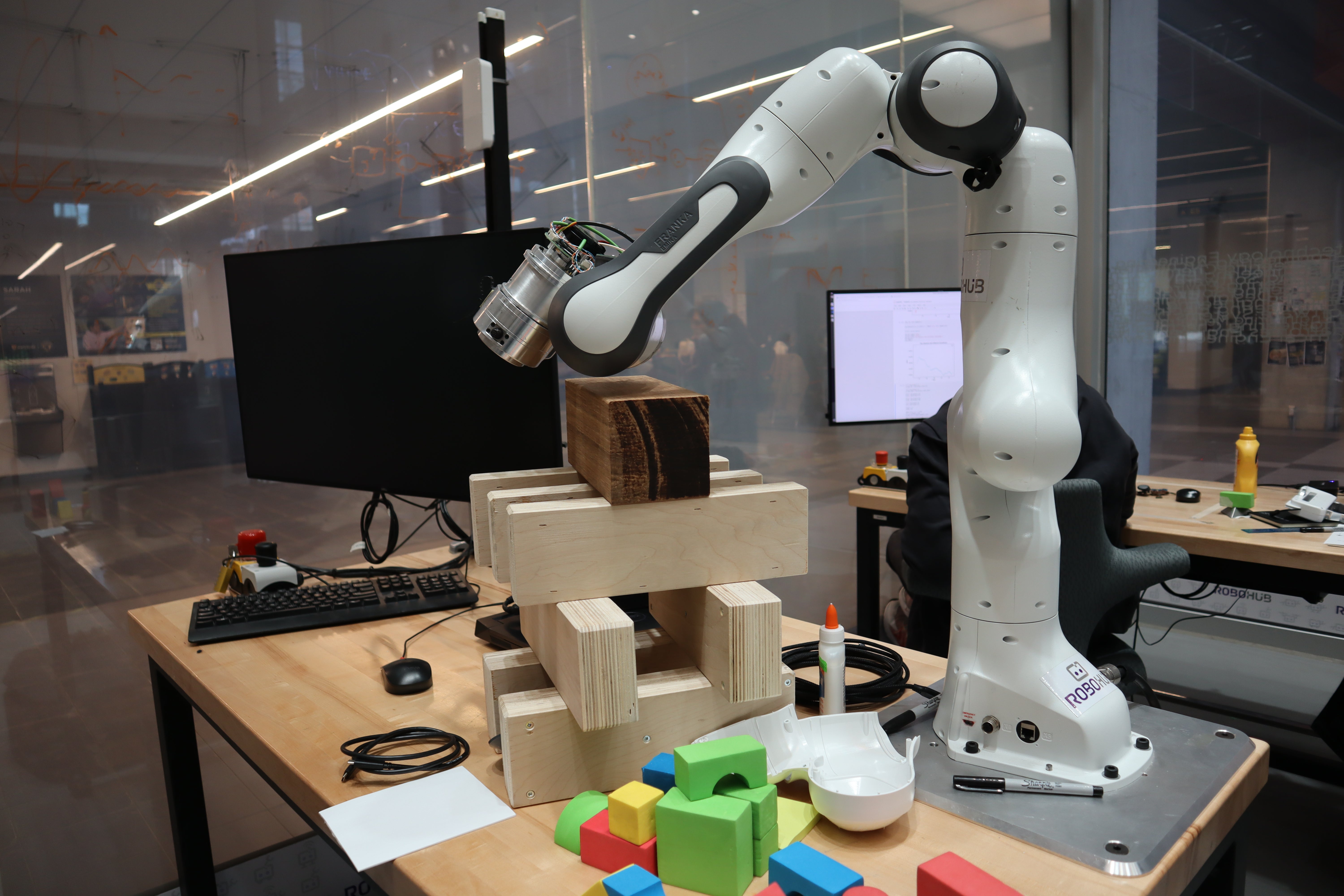 A robotic arm is positioned over a stack of wooden blocks on a table. There are colorful building blocks scattered on the table, and a computer monitor is visible in the background.