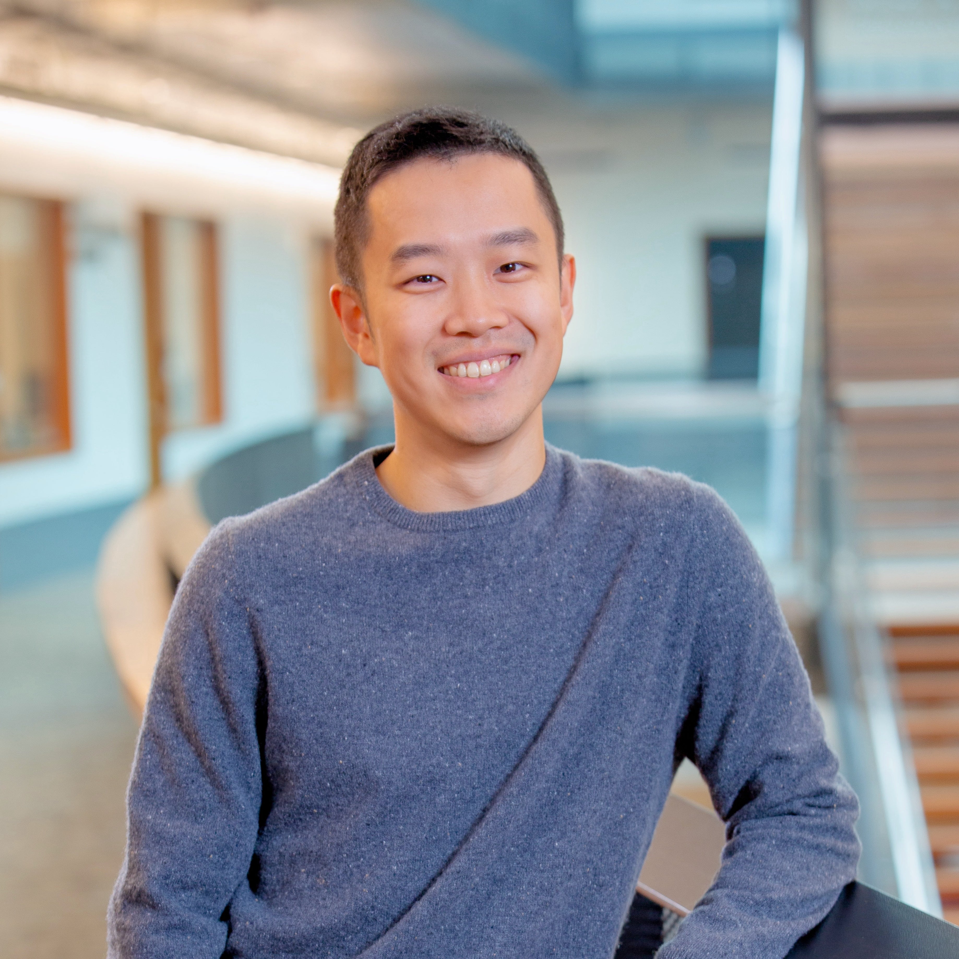 An Asian man donning a grey-blue long sleeve shirt posing and smiling in front of a staircase.