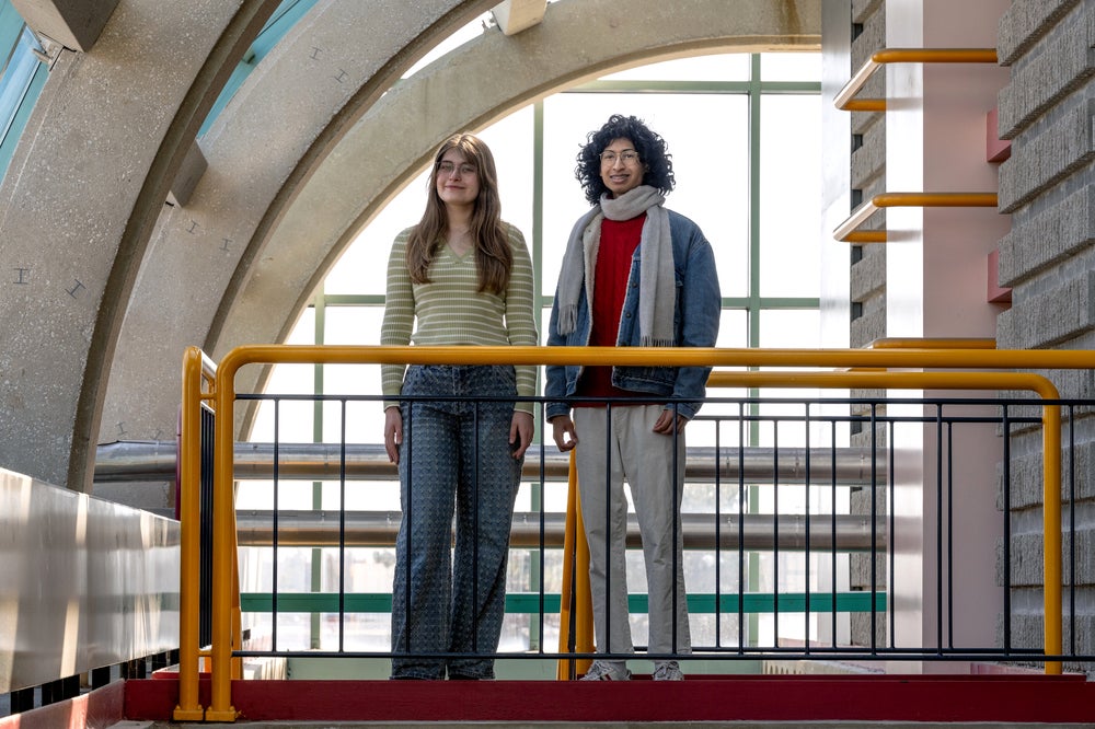 Two students standing on a podium in front of a yellow bar. The left is a blonde woman wearing glasses and a light-green long sleeve shirt and jeans. left is a south asian man with fluffy hair, wearing a grey scarf, red shirt and a demin jacket