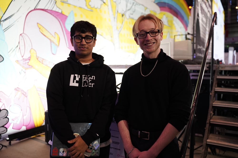 Two men both wearing glasses (left south asian, right is a blond Caucasian) posing with their hands enclosed in front of them. The person on the left is holding a laptop