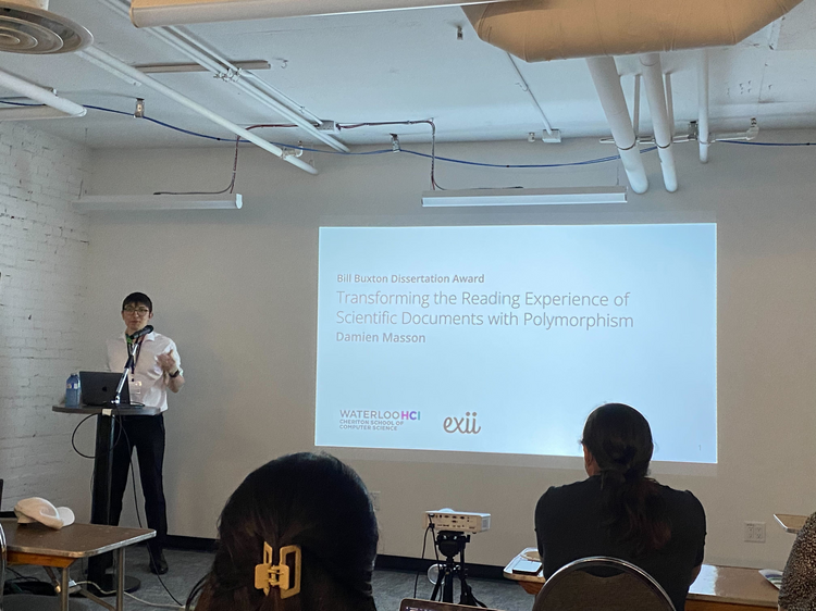 Photo of Damien at GI 2024, presenting with a slideshow to a crowd
