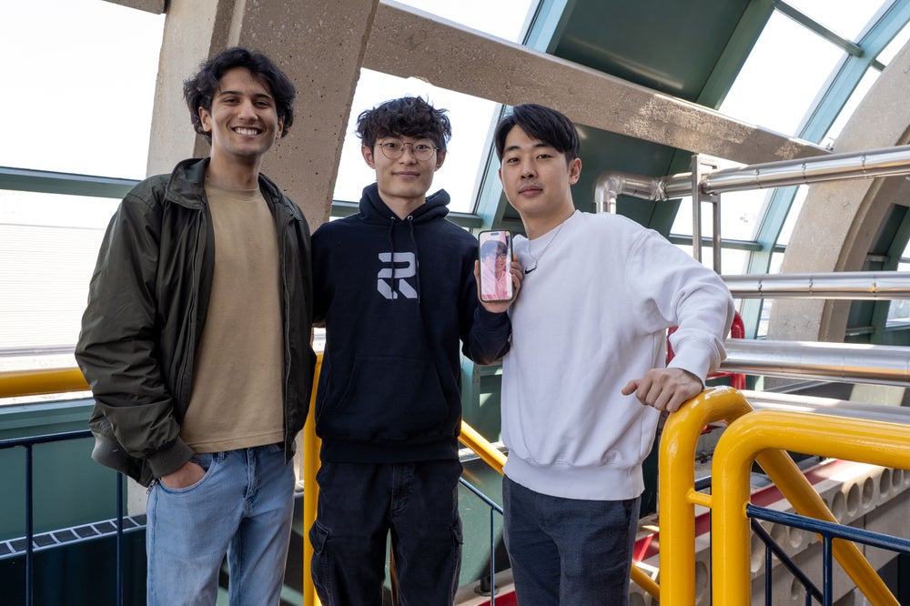 Photo of three men in casual clothing. The man in the middle is holding a phone that displays a photo of their teammate, who wasn't available to be photographed