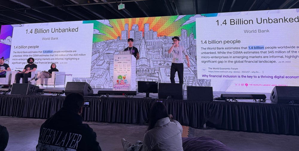 Two asian men standing on podium. Their background has a slidedeck (showing on 2 screens) with text saying "1.4 Billion Unbanked". In between the screen is a black and white city landscape, while the sky has red, green, orange, blue streaks.